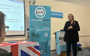 Victoria Carson standing at the front of a hall, smiling and speaking as she welcomes Reform UK members and supporters to a local engagement event in Battle on 31 October 2025.
